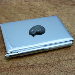 Könnte beinhalten: Ein silberner Kartenhalter aus Metall mit einer kleinen Igelfigur auf der Vorderseite. Der Halter ist geschlossen und hat ein silbernes Scharnier.