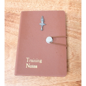 Op de afbeelding: Een bruin leren notitieboekje met een zilveren ballerina-bedel en de woorden "Training Notes" in goud gedrukt.