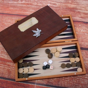 Könnte beinhalten: Ein Backgammon-Spiel aus Holz mit einem braunen Lederetui. Das Etui hat eine Messingplakette mit einer Inschrift. Das Spielbrett ist geöffnet und zeigt die Spielfläche mit schwarzen und weißen Feldern. Auf dem Brett befinden sich weiße und braune Spielsteine.