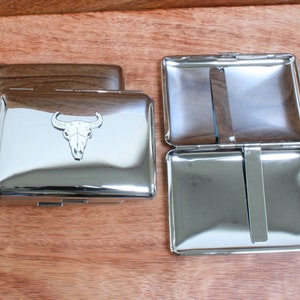 Op de afbeelding: Twee rechthoekige metalen kaarthouders. Eén is gesloten met een houten bovenkant, de andere is open. De gesloten houder heeft een zilveren afwerking met een schedelontwerp van een stier. De open houder heeft een zilveren afwerking met een metalen clip aan de binnenkant.