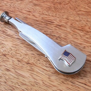 May include: A silver pipe tool with a small American flag on the side. The tool has a built-in tamper and a small blade for cutting tobacco.