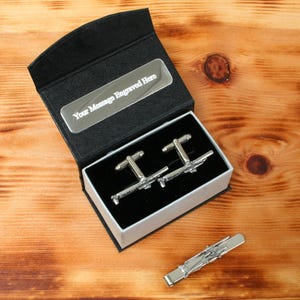 Könnte beinhalten: Eine schwarze Geschenkbox mit einem silbernen Flugzeug-Manschettenknopf-Set und einer passenden Krawattenklammer. Die Box hat eine silberne Plakette mit der Aufschrift "Your Message Engraved Here".