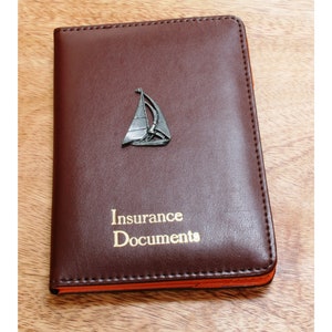 May include: A brown leather insurance document holder with a gold inscription that reads "Insurance Documents". A small silver sailboat is attached to the front of the holder.