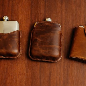 Op de afbeelding: Drie bruine leren heupflessen met zilveren doppen liggen op een houten oppervlak. De flessen hebben verschillende ontwerpen, waaronder een met een leren hoes, een met getextureerd leer en een met een klepsluiting.