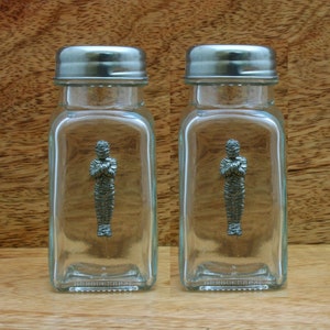 May include: A set of two glass salt and pepper shakers with silver lids. Each shaker features a pewter figurine of a mummy inside.
