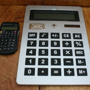 Puede incluir: Una calculadora plateada con botones negros y una pequeña calculadora negra a su lado. La calculadora más grande tiene una placa de metal plateada con el texto "Your Message Engraved Here For FREE".