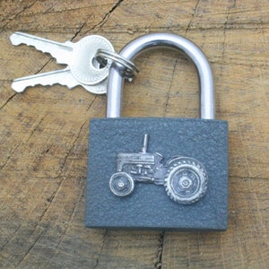 May include: A gray padlock with a silver tractor design on the front. The padlock has a silver shackle and is attached to two silver keys.