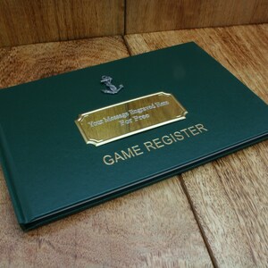 Puede incluir: Un libro encuadernado en cuero verde con una placa dorada que dice "Your Message Engraved Here For Free" y "GAME REGISTER". Un pequeño ancla de plata está unida a la portada.