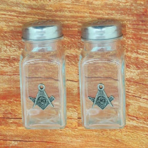 May include: Two clear glass salt and pepper shakers with silver lids. Each shaker features a silver Masonic symbol.