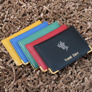 May include: A stack of colorful travel card holders in yellow, blue, green, red, and black. The black holder has a silver emblem and the words "Travel Card" in gold. Each holder has gold corner accents.