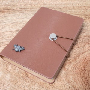 May include: A brown leather-bound journal with a textured cover. The journal features a metal bee embellishment and a button closure with an elastic band. The journal is closed and resting on a wooden surface.