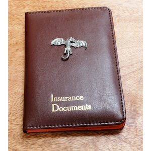 May include: A brown leather insurance document holder with a silver dragon design and the words "Insurance Documents" in gold lettering.