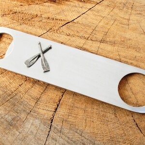 Puede incluir: Un abridor de botellas plateado con un diseño de remos cruzados. El abridor está sobre una superficie de madera.