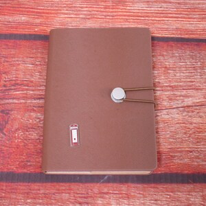 May include: A brown leather journal with a silver button closure and a brown elastic band. A small red and white telephone booth emblem is on the front cover. The journal is closed and resting on a wooden surface.