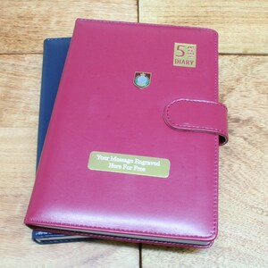 May include: A red leather 5 year diary with a gold inscription that says "5 Year Diary" and a gold plaque that says "Your Message Engraved Here For Free".
