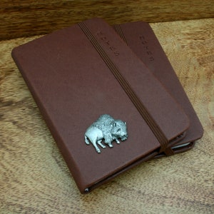 May include: Two brown leather notebooks with the word "NOTES" embossed on the cover. A silver-colored bison charm is affixed to the front of one notebook. The notebooks are bound with a brown elastic band.