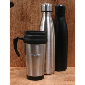 Könnte beinhalten: Ein Edelstahl-Reisebecher mit schwarzem Griff und Deckel sowie zwei Edelstahl-Wasserflaschen, eine silberne und eine schwarze, werden auf einer Holzoberfläche präsentiert. Der Reisebecher hat ein kleines Flugzeugemblem.