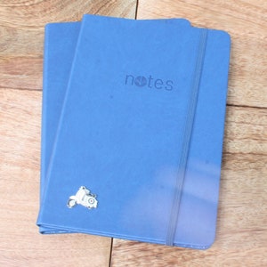 May include: Two blue notebooks with a black elastic band closure. The top notebook has the word "notes" printed in white on the front cover. A silver scooter charm is attached to the bottom notebook.