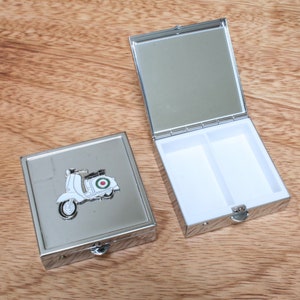 Könnte beinhalten: Silberne Pillendose mit einem Scharnierdeckel und zwei Fächern. Die Vorderseite der Dose zeigt einen silbernen Roller mit einem grün-weißen Zielscheiben-Design.