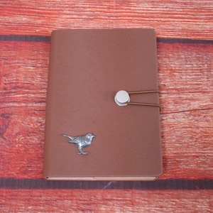 May include: A brown leather journal with a silver bird design and a silver button closure. The journal is on a red wooden background.