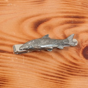 Könnte beinhalten: Ein silberner Krawattenclip mit einem detaillierten Fischdesign. Der Fisch blickt nach links und hat eine realistische Darstellung.