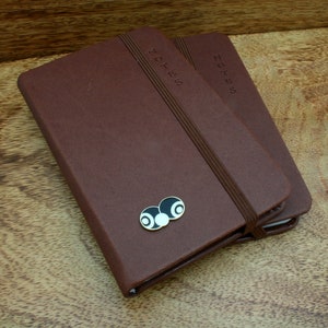 May include: Two brown leather bound notebooks with elastic closures. The top notebook has a black and white enamel pin of two billiard balls.