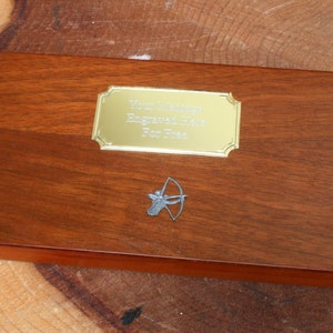 Może przedstawiać: Drewniana skrzynka z pozłacaną tabliczką z napisem "Ręcznie grawerowany nóż dla Ciebie" i srebrną figurką łucznika.