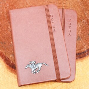May include: Two brown leather-bound notebooks with the word "NOTES" embossed on the front. The notebook on the left has a silver metal horse and jockey design.