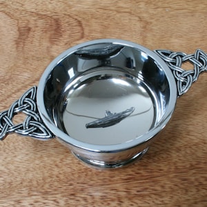 Puede incluir: Una taza de plata con un asa de nudo celta y un diseño de submarino en el fondo.