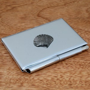 Op de afbeelding: Een zilveren metalen kaarthouder met een schelpontwerp. De houder heeft een scharnierende klep en een zilveren metalen clip.