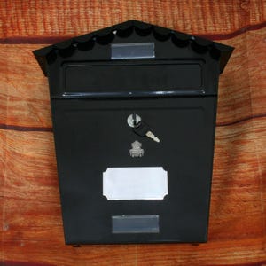 Könnte beinhalten: Schwarzer Metallbriefkasten mit einem Schlitz für die Post, einem Schlüsselloch und einem leeren Namensschild. Der Briefkasten hat ein dekoratives, gewelltes Oberteil.
