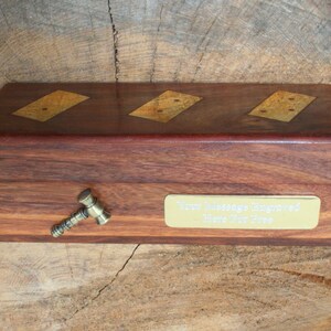 May include: A wooden box with a brass inlay design. The box has a brass plaque on the front that says "Your Message Engraved Here For Free". A small brass hammer is attached to the side of the box.