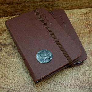 Puede incluir: Dos cuadernos encuadernados en cuero marrón con cierres elásticos. El cuaderno superior tiene un emblema circular de metal con un lobo aullando a la luna.