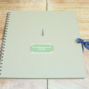 May include: A brown spiral bound scrapbook with a green label that says "Your Message Engraved Here For Free". A blue ribbon is tied around the spine of the book.