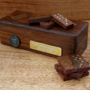 Puede incluir: Un juego de dominó de madera con una caja de madera personalizada. La caja tiene una placa de latón con el texto "Su mensaje grabado aquí gratis". Los dominós están hechos de madera oscura con puntos de latón.