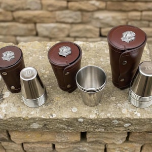 Könnte beinhalten: Ein Set aus braunen Leder-Würfelbechern mit silbernen Akzenten und einem Metall-Hundeemblem. Das Set enthält Edelstahl-Würfelbecher mit der Zahl 10. Auf einer Steinoberfläche angeordnet.