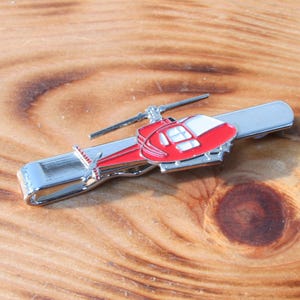 Könnte beinhalten: Ein silberner Krawattenclip mit einem roten und weißen Hubschrauber-Design. Der Hubschrauber hat einen silbernen Propeller und eine weiße Windschutzscheibe.