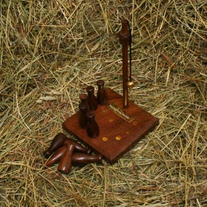 Peut inclure: Un jeu de table en bois avec une base carrée, un poteau vertical avec une boule suspendue et de petites quilles en bois. Le jeu est présenté sur un lit de foin, soulignant le savoir-faire artisanal.