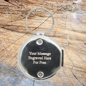Op de afbeelding: Een zilveren intrekbare sleutelhanger met een rond metalen label. Het label heeft de tekst "Your Message Engraved Here For Free" gegraveerd.
