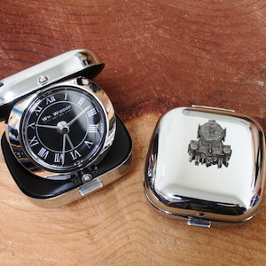 Puede incluir: Un reloj de bolsillo plateado con una esfera negra y números romanos. El reloj está en una caja plateada con una tapa con bisagras. La caja tiene un pequeño emblema plateado de una locomotora en la parte delantera.