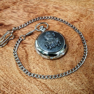May include: A silver pocket watch with a chain. The watch face features a detailed engraving of a steam locomotive.