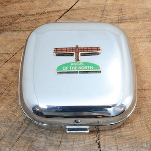 May include: A silver metal box with a hinged lid. The box has a small, metal pin attached to the lid. The pin features a brown and gold sculpture of the Angel of the North, a famous landmark in Gateshead, England. The pin also has a green sign that reads "Angel of the North".