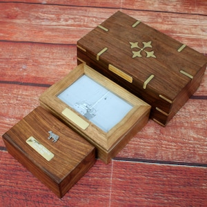 Può includere: Tre scatole di legno con dettagli in ottone. La scatola più grande ha un design decorativo in ottone sul coperchio. La scatola centrale ha una cornice per foto con un'immagine in bianco e nero di una barca a vela. La scatola più piccola ha una statuetta di mucca in ottone sul coperchio.