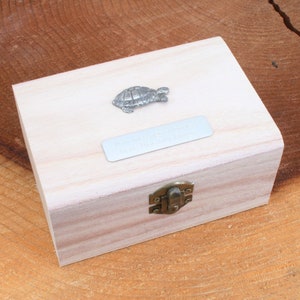 Puede incluir: Una pequeña caja de madera con tapa abatible y cierre de metal. La caja tiene una figurita de tortuga plateada en la parte superior y una placa plateada con una inscripción.