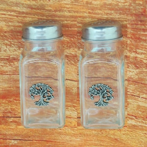 May include: Two clear glass salt and pepper shakers with silver metal lids. Each shaker has a decorative silver tree emblem on the front. The shakers are rectangular and sit on a wooden surface.