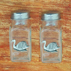 Pode incluir: Dois saleiros e pimenteiros de vidro com tampas prateadas. Cada saleiro tem um design de cisne de peltre.