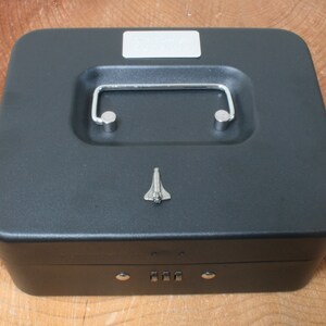 May include: A black metal cash box with a combination lock and a silver handle. The box has a small silver space shuttle design on the top.