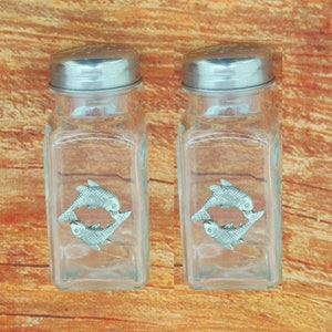 Könnte beinhalten: Zwei klare Glassalz- und Pfefferstreuer mit silbernen Deckeln. Jeder Streuer ist mit einem Zinnfisch-Design verziert.