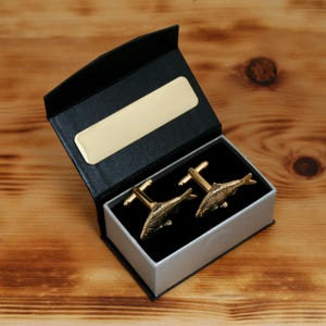 May include: Gold-toned fish cufflinks presented in a black and gray box with a gold-colored nameplate. The cufflinks feature a detailed fish design. The box is open, revealing the cufflinks against a black velvet interior, set on a wooden surface.