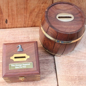 May include: Two wooden money boxes. One is a barrel shape with brass bands and the other is a square box with a hinged lid. Both have a slot for coins and a small metal ornament on top.